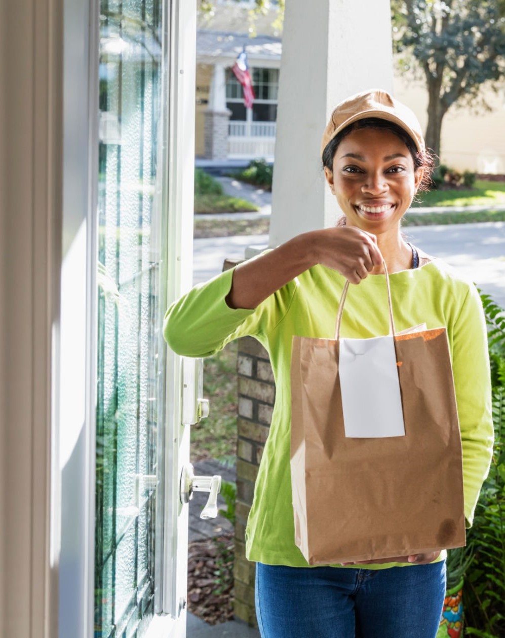 A young African-American woman in her 20s standing outside the front door of a home, looking at the camera and holding a bag of take out food ordered from a restaurant. She could be the delivery person, or perhaps she is the homeowner who has picked up her own